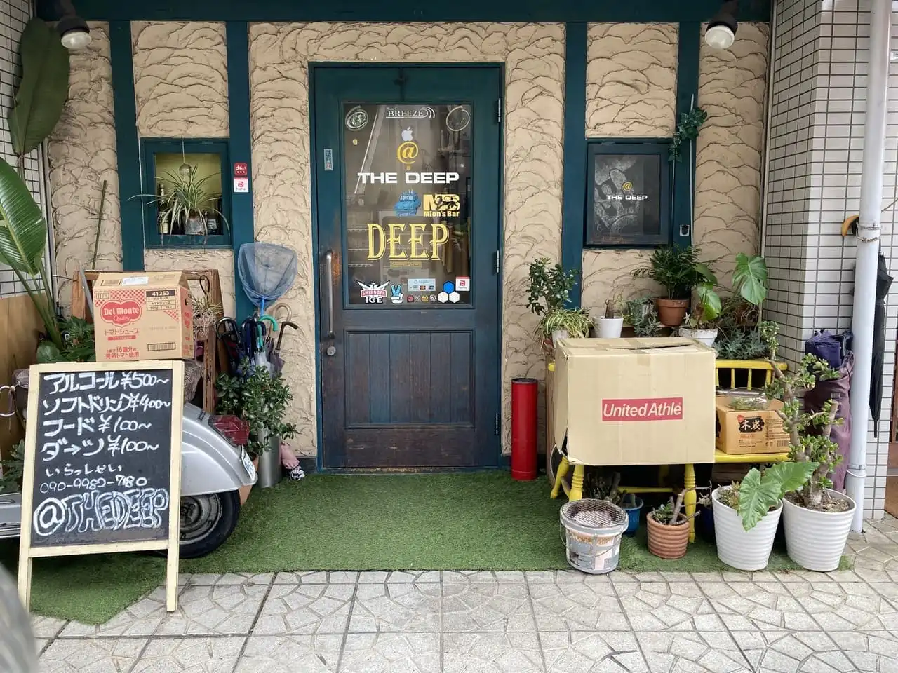 奈良市 法華寺町のbar @ the deepの外観写真です。緑の扉のガラスには@ the deepの文字、ステッカーなどが貼られています。お店の前には観葉植物や傘立て、バイク、看板などが置かれています。看板にはあるコール￥500～、ソフトドリンク￥400～、フード￥100円～、ダーツ￥100～、いらっしゃい090-9982-7180、@thedeepと書かれています。