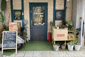 奈良市 法華寺町のbar @ the deepの外観写真です。緑の扉のガラスには@ the deepの文字、ステッカーなどが貼られています。お店の前には観葉植物や傘立て、バイク、看板などが置かれています。看板にはあるコール￥500～、ソフトドリンク￥400～、フード￥100円～、ダーツ￥100～、いらっしゃい090-9982-7180、@thedeepと書かれています。