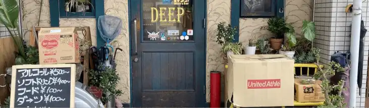 奈良市 法華寺町のbar @ the deepの外観写真です。緑の扉のガラスには@ the deepの文字、ステッカーなどが貼られています。お店の前には観葉植物や傘立て、バイク、看板などが置かれています。看板にはあるコール￥500～、ソフトドリンク￥400～、フード￥100円～、ダーツ￥100～、いらっしゃい090-9982-7180、@thedeepと書かれています。