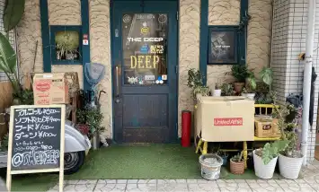 奈良市 法華寺町のbar @ the deepの外観写真です。緑の扉のガラスには@ the deepの文字、ステッカーなどが貼られています。お店の前には観葉植物や傘立て、バイク、看板などが置かれています。看板にはあるコール￥500～、ソフトドリンク￥400～、フード￥100円～、ダーツ￥100～、いらっしゃい090-9982-7180、@thedeepと書かれています。