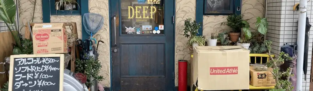 奈良市 法華寺町のbar @ the deepの外観写真です。緑の扉のガラスには@ the deepの文字、ステッカーなどが貼られています。お店の前には観葉植物や傘立て、バイク、看板などが置かれています。看板にはあるコール￥500～、ソフトドリンク￥400～、フード￥100円～、ダーツ￥100～、いらっしゃい090-9982-7180、@thedeepと書かれています。